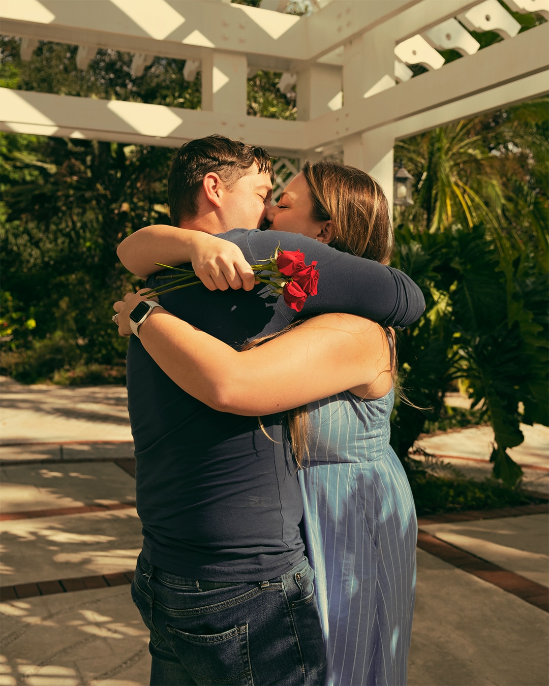 man and woman hug while woman holds red flowers under white gazebo type structure outside in garden