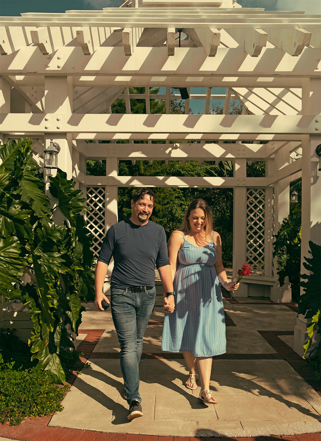 man and woman hold hands while woman holds red flowers in hand as they walk away from under white gazebo structure