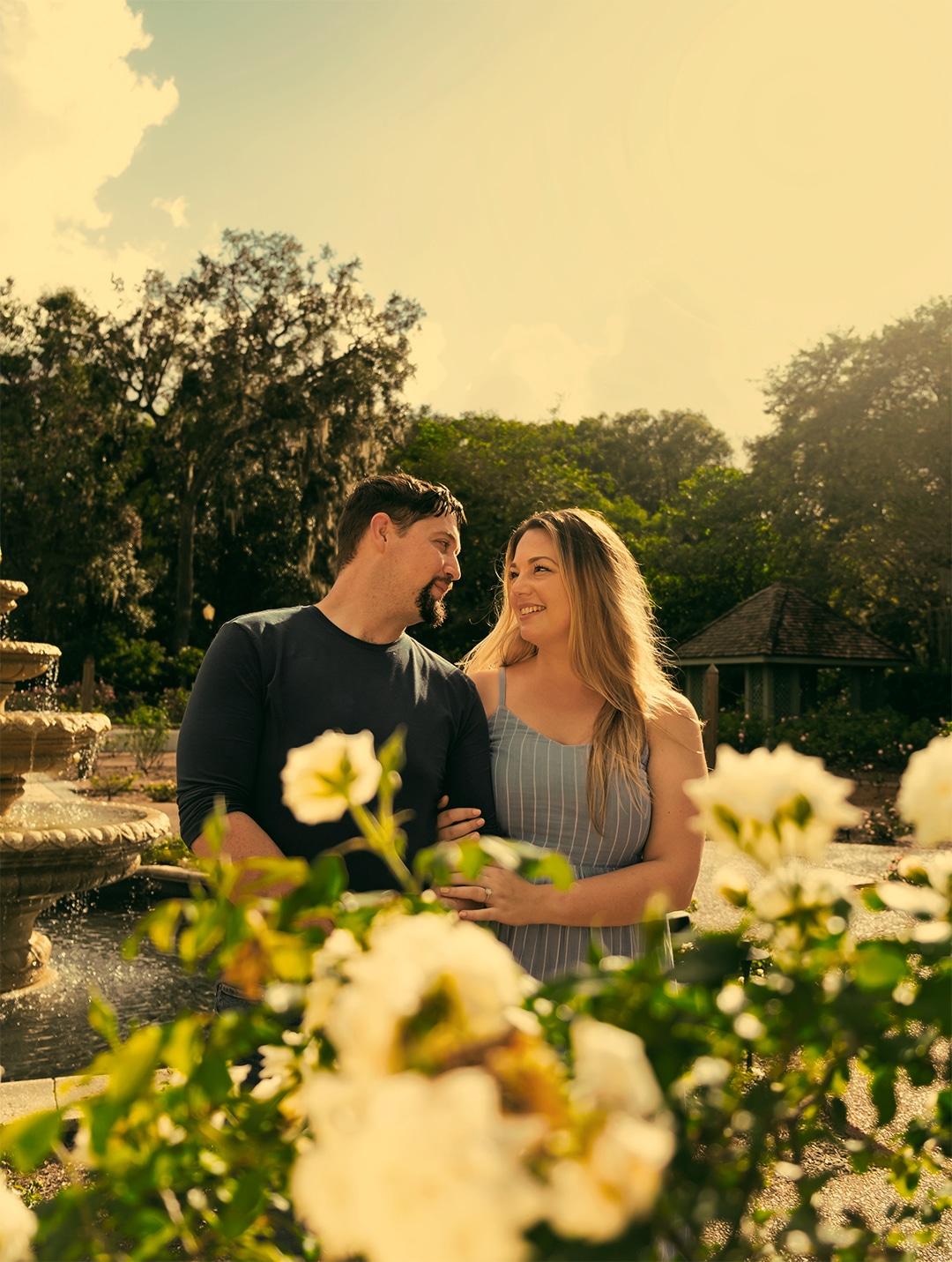 man and woman look at each other outside in gardens with flowers in front of them and fountain to the left