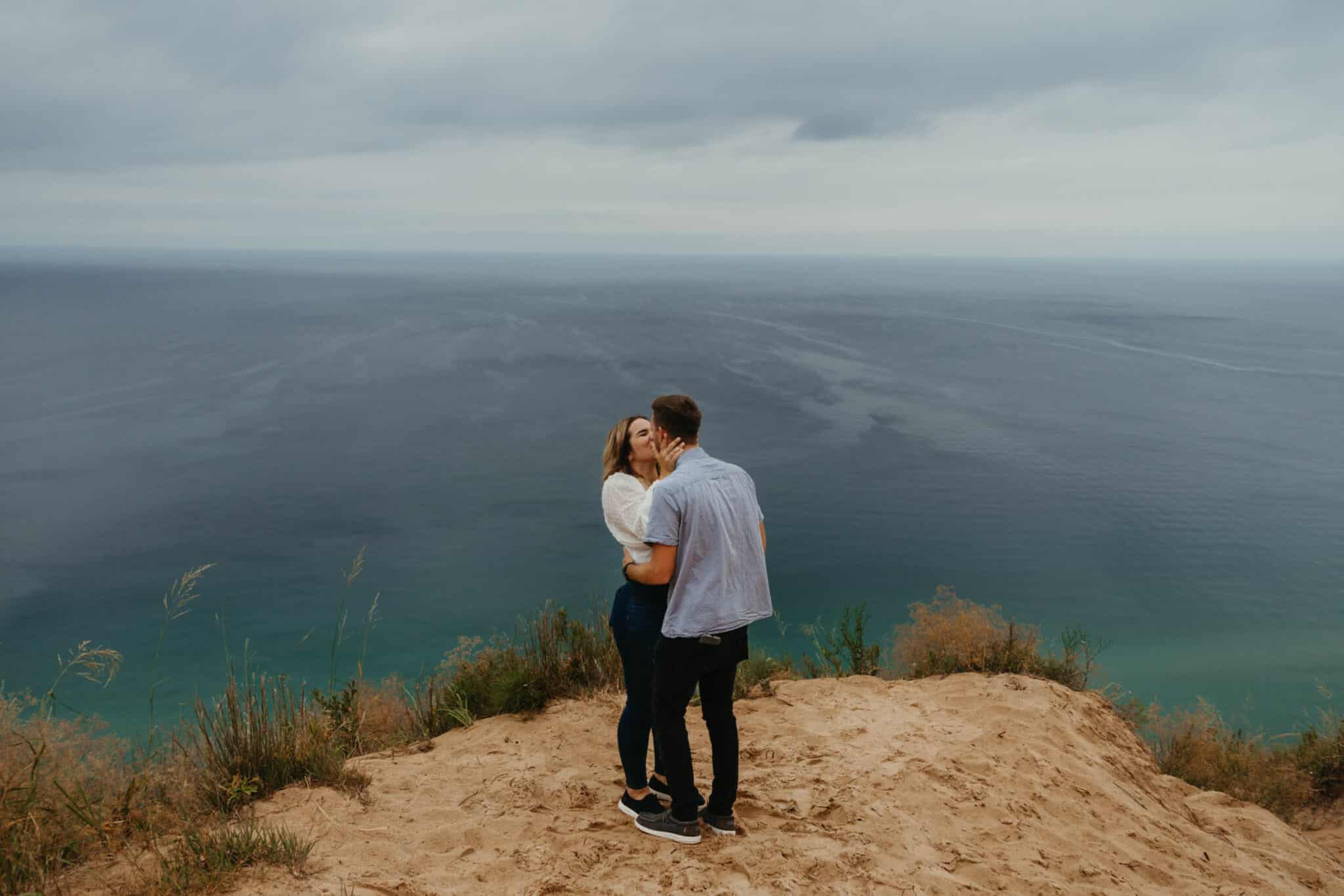 man and woman stand outside on elevated piece of land overlooking giant body of water kissing each other