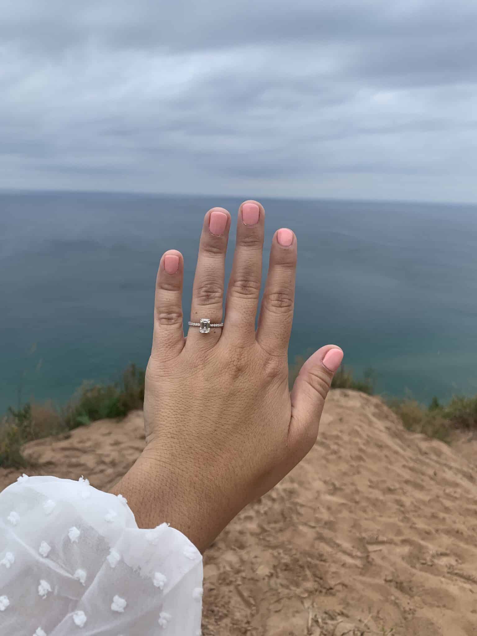 woman's hand outstretched in front of land and body of water with pink nail polish showing off new engagement ring
