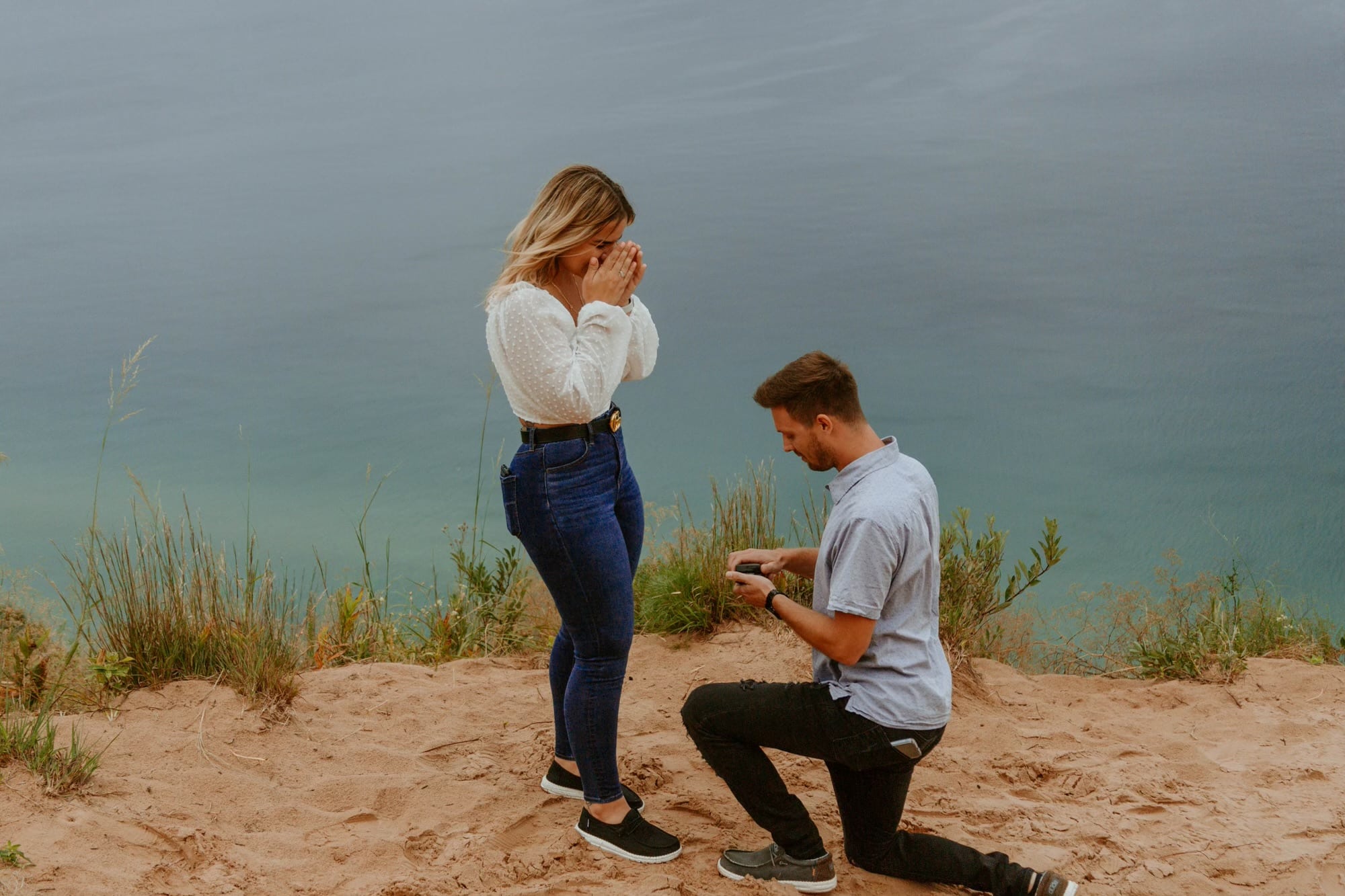 man kneels down on the ground in traverse city marriage proposal while woman stands shocked in front of him