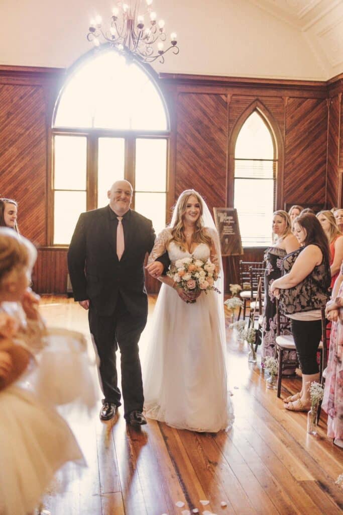 father of the bride walking her down the aisle with sunlight streaming through window behind them at THE 1883 Historic Venue