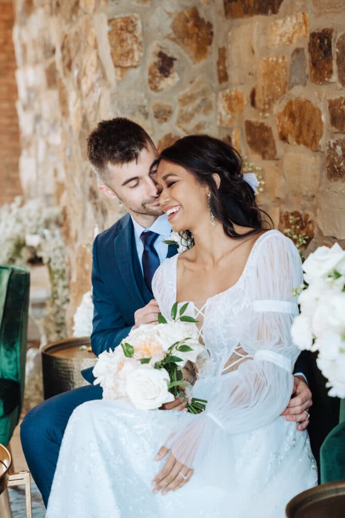 photo by Bouquet Photography of bride and groom sitting against a stone wall smiling and laughing with each other