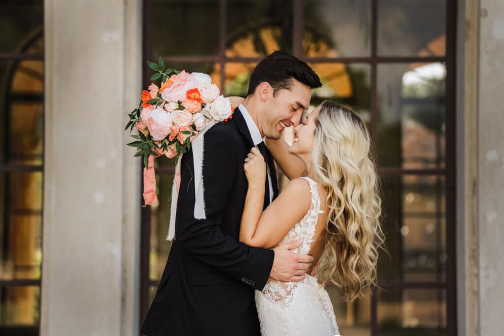 bride with bouquet of pink and salmon, and groom kissing in front of glass wall, photo by Taylor Kuperberg Photography