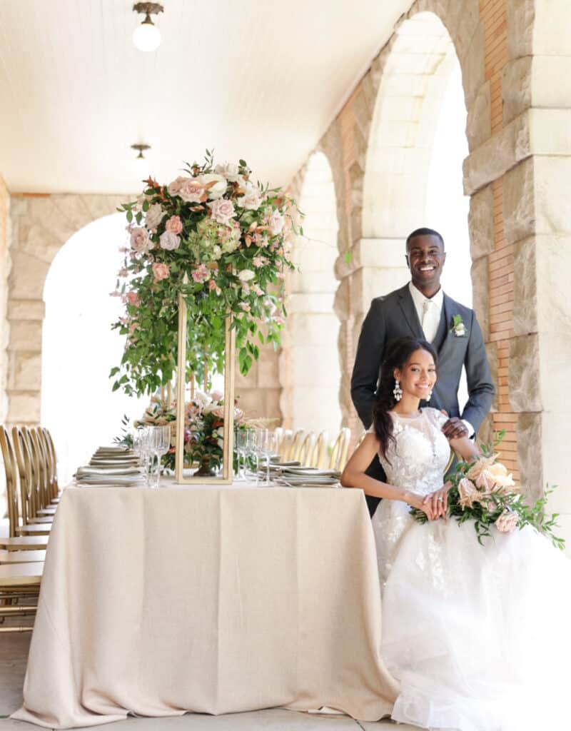 photo of bride sitting and groom standing behind her at reception table with tall centerpiece in covered alcove by Sylvia Santos Photography