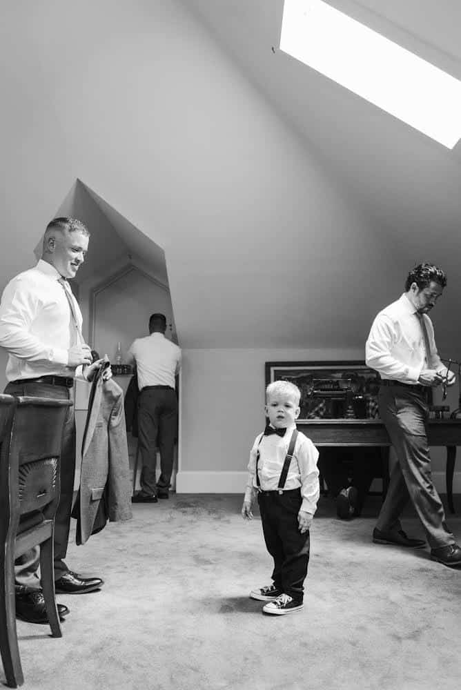 black and white photo of ring bearer getting ready with groomsmen under eaves of building at THE 1883 Historic Venue
