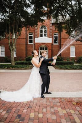 photo by Taylor Kuperberg Photography of groom standing on bricked street spraying champagne and bride holding on to his shoulders standing behind him