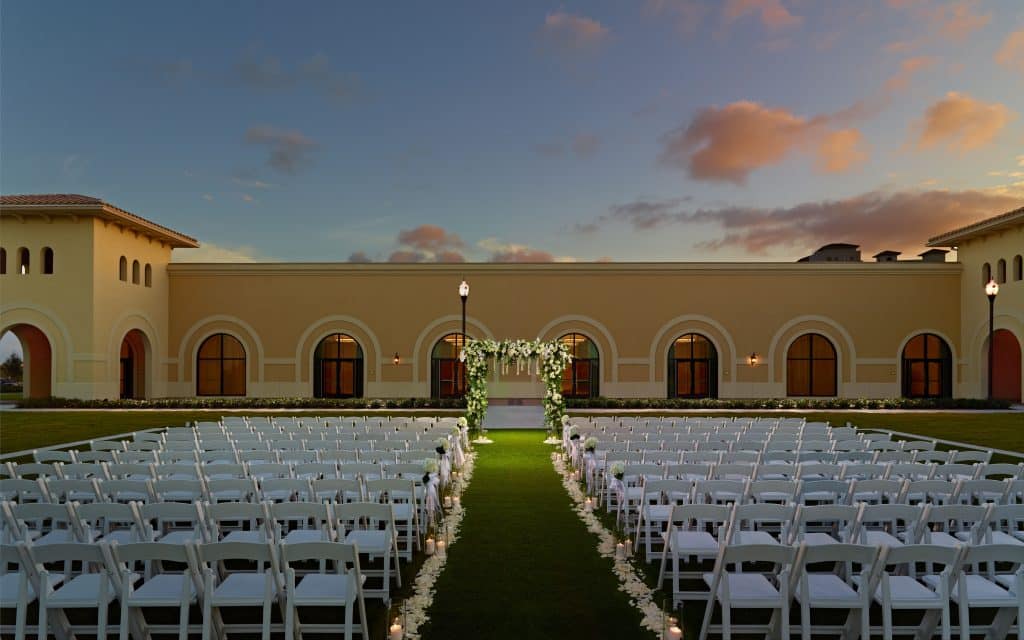 long grass aisle lined with uplights and white chairs for outdoor ceremony leading toward long arched wall at sunset at Omni Orlando Resort at Champions Gate