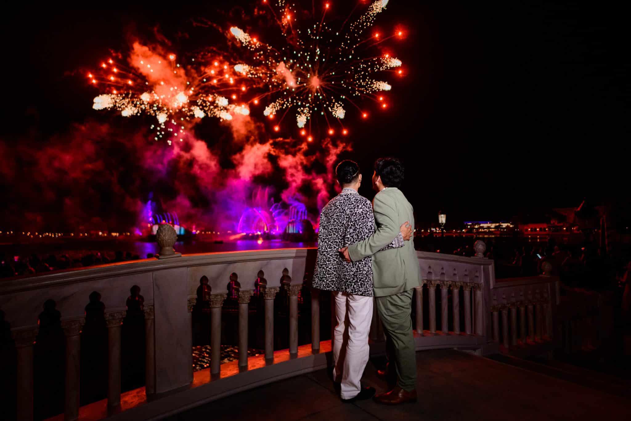 Couple faces a waterfront view as fireworks go off in the distance