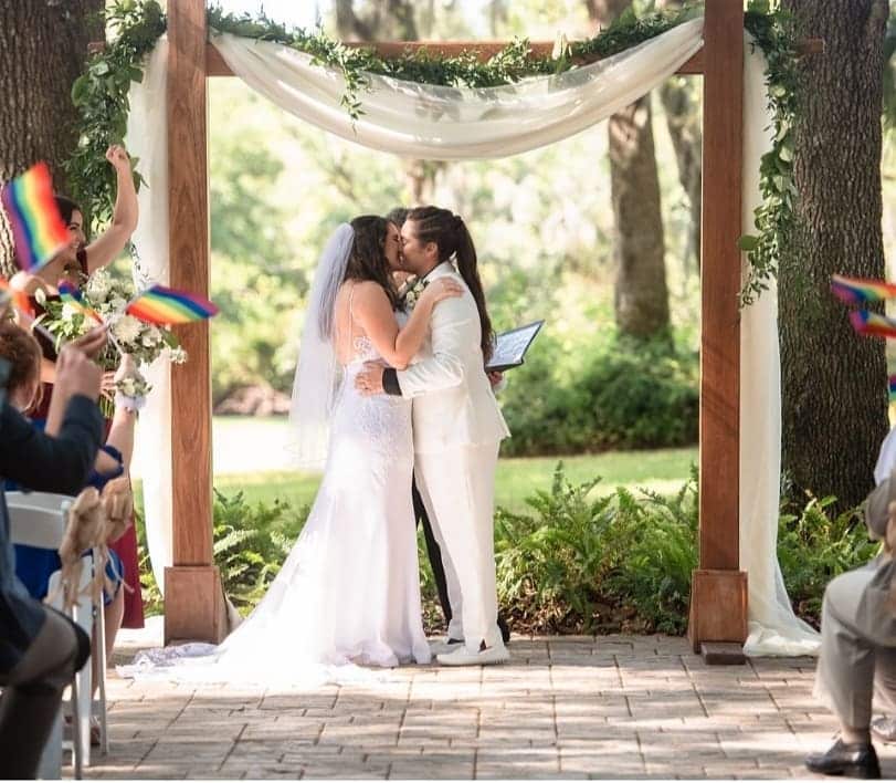 brides kissing under arbor swagged in white and green with rainbow flags waved by wedding guests coordinated by Julie Miner Events