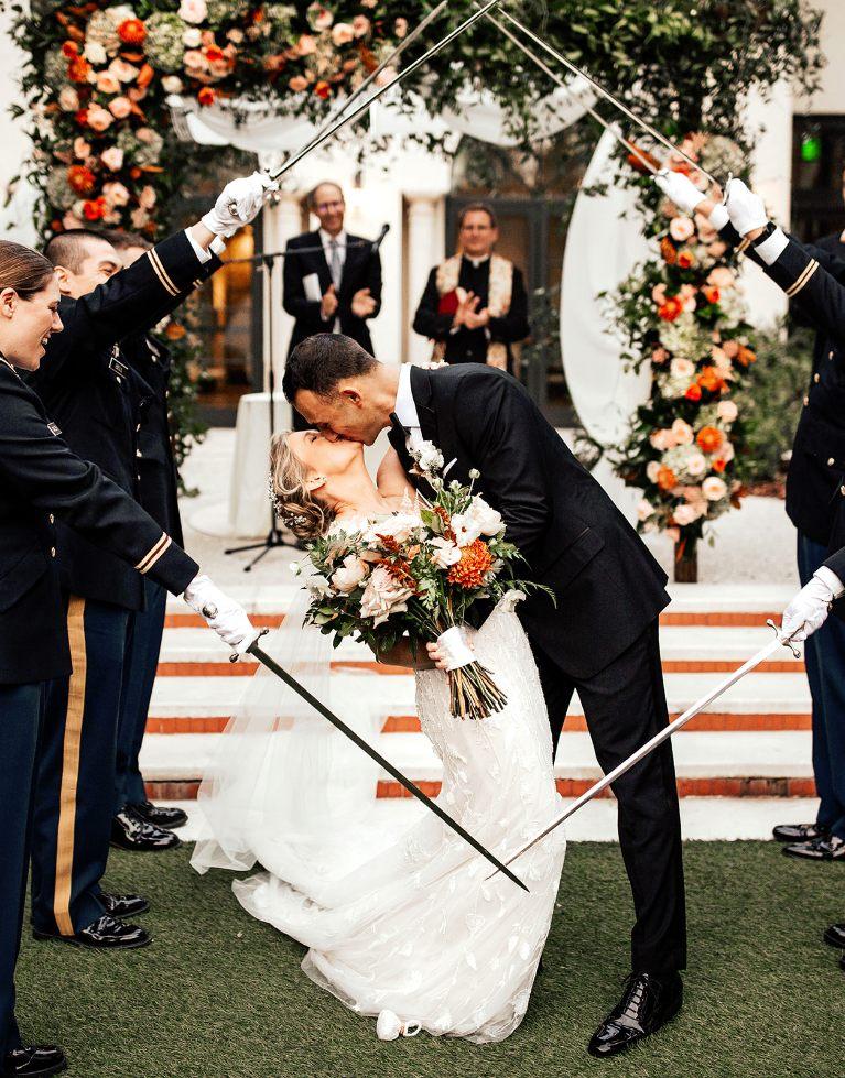 groom dipping bride holding bouquet of pink and green flowers under arch of matching flowers and greenery by Ian Tafoya Designs in military arch of ceremonial swords