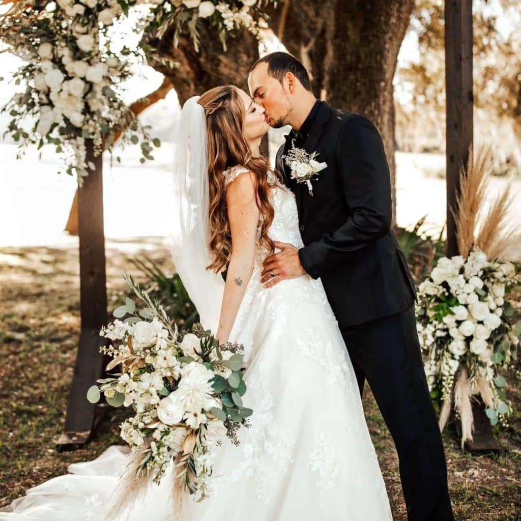 bride holding large bouquet of green and white flowers by Ian Tafoya Designs kissing groom among trees with flowering plants