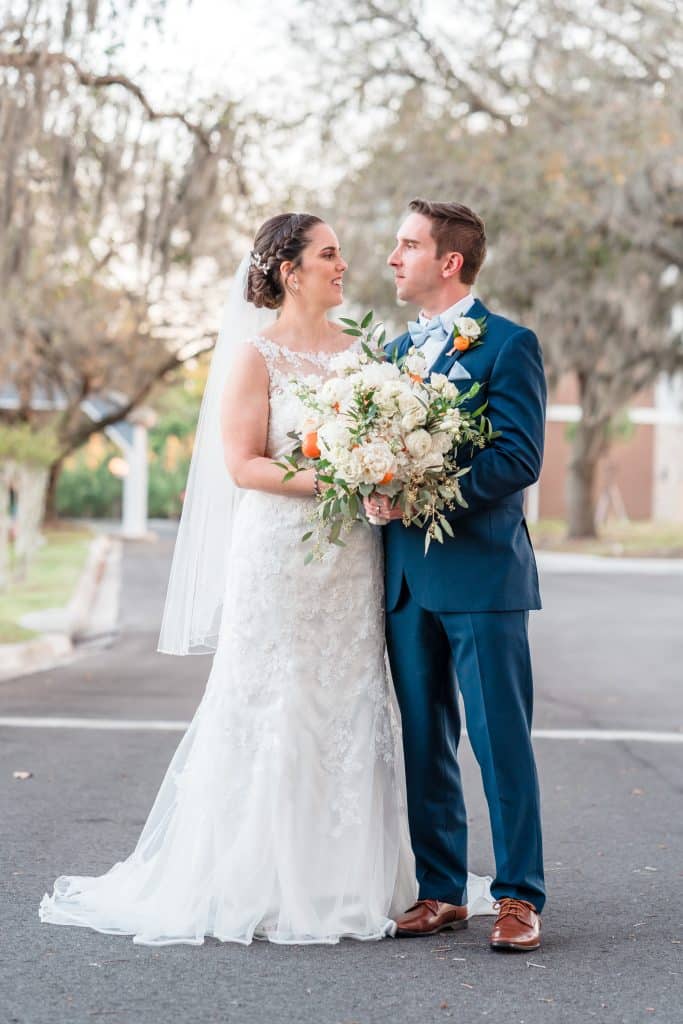 bride and groom wearing wedding attire from Carolyn Allen’s Bridals & Formals standing in the middle of a tree lined street holding large bouquet of pink and white flowers