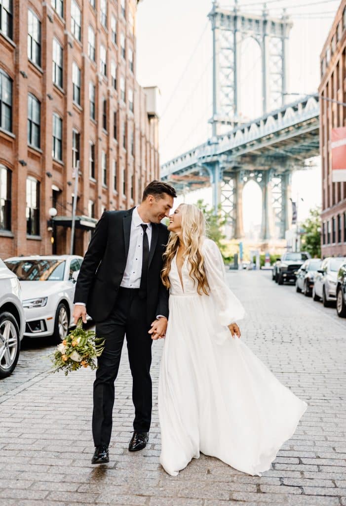 bride and groom standing in street kissing underneath the Brooklyn Bridge wearing wedding gown and suit from Carolyn Allen’s Bridals & Formals