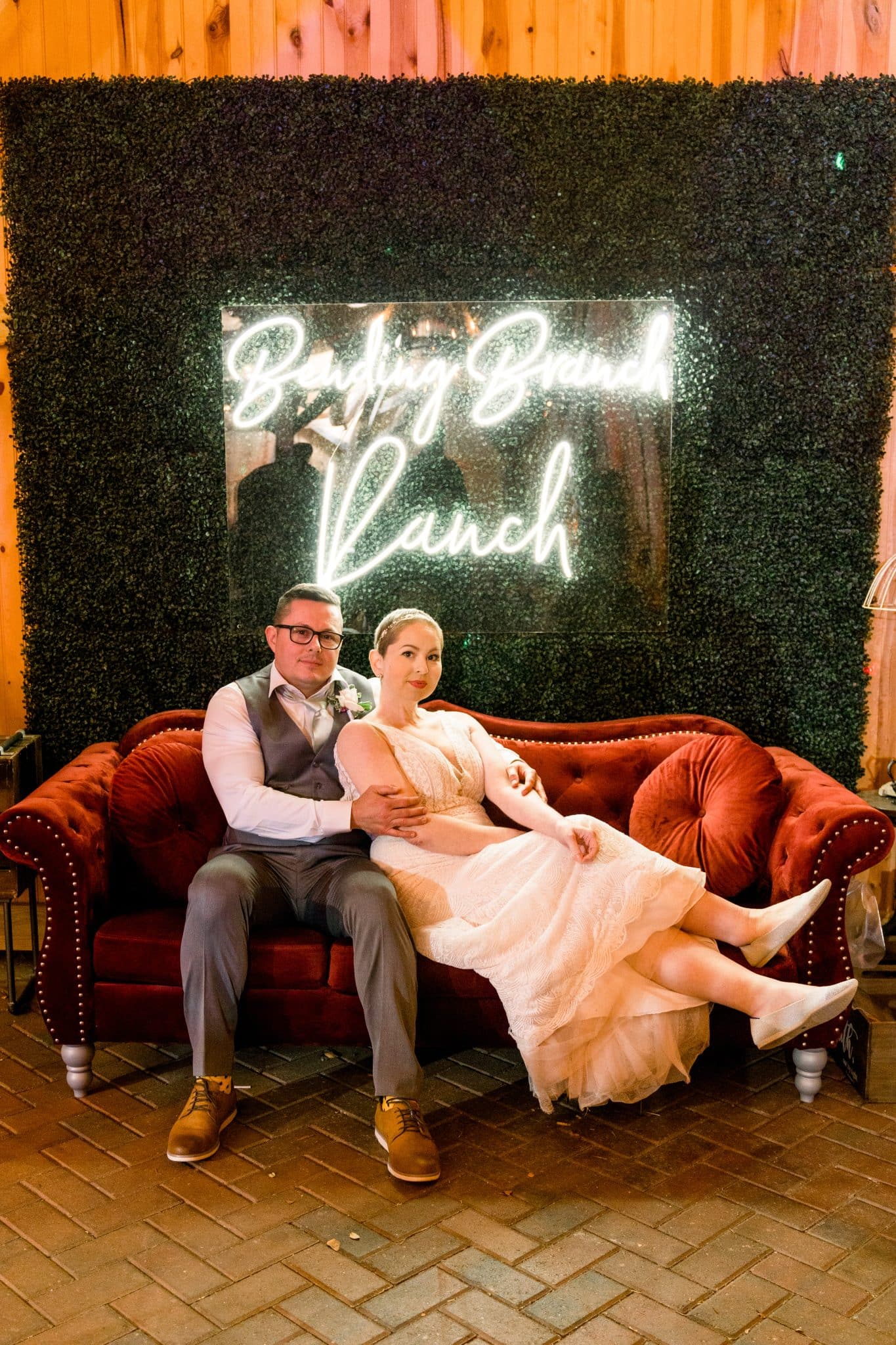 Bride and groom sit on a couch together in front of a light up "Bending Branch Ranch" sign