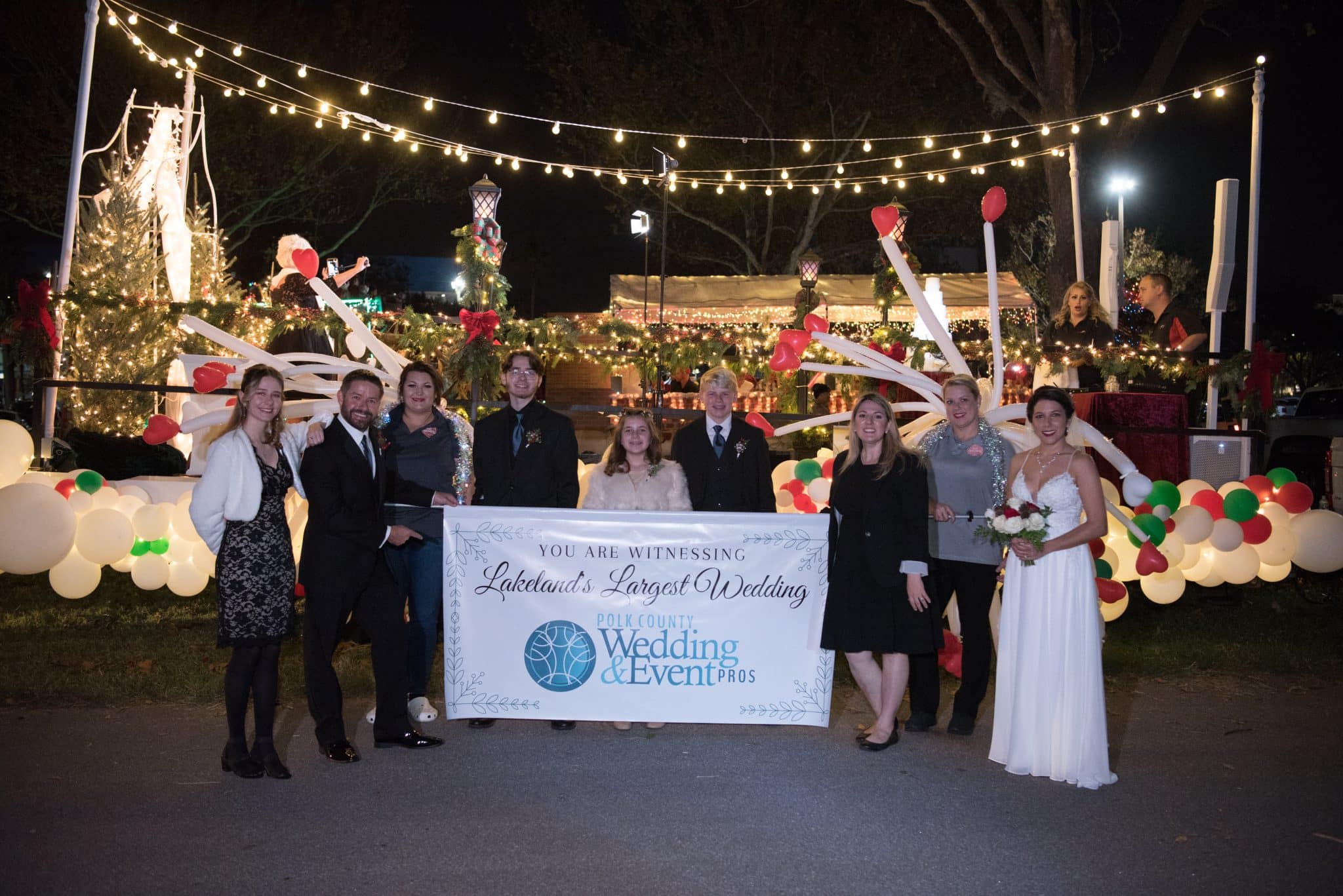 Wedding party gathers in front of Christmas themed parade float holding a sign reading "Lakeland's Largest Wedding by Polk County Wedding & Events Pros"