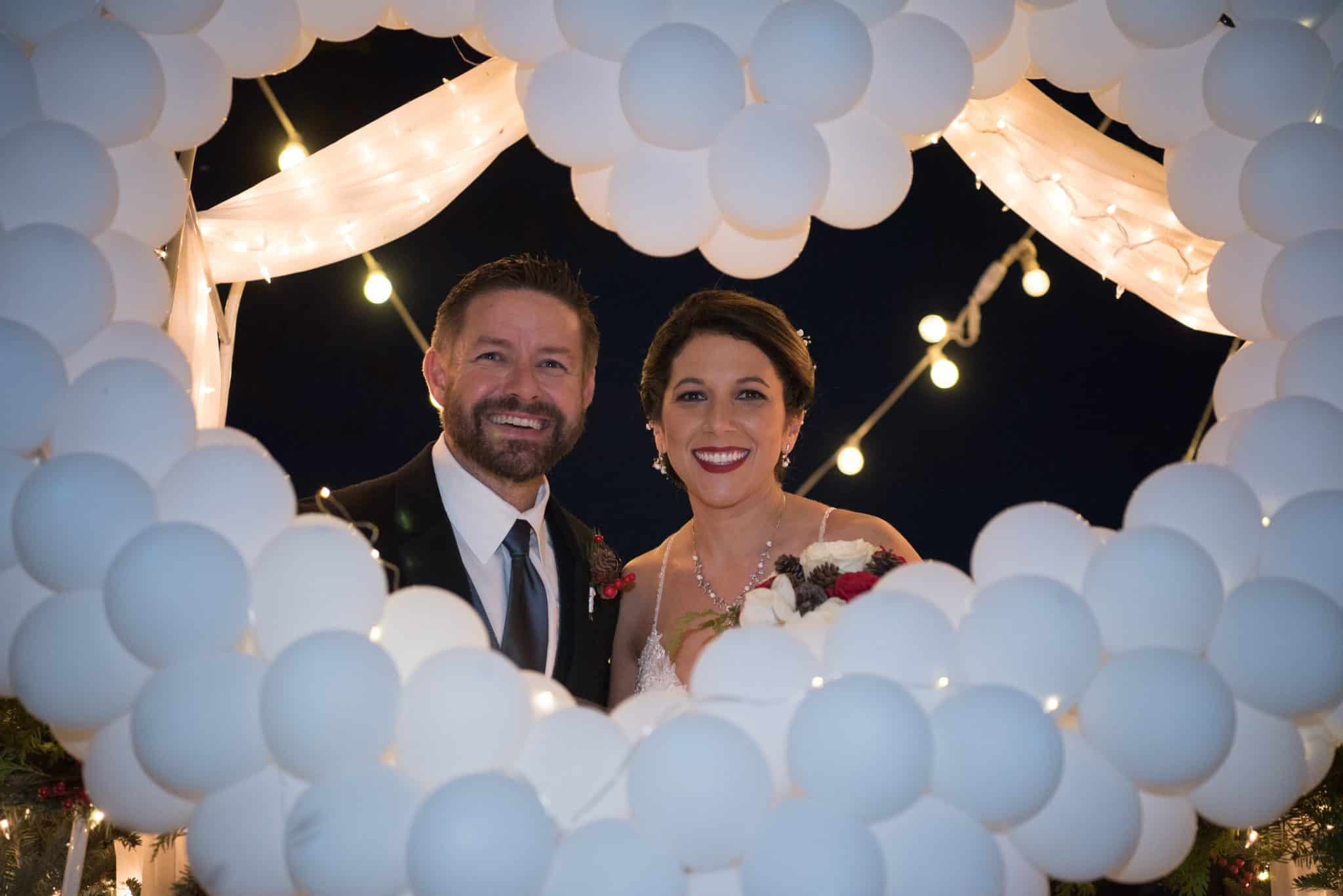 Newlywed couple poses for a picture with dozens of white balloons in the shape of a heart framing them. 
