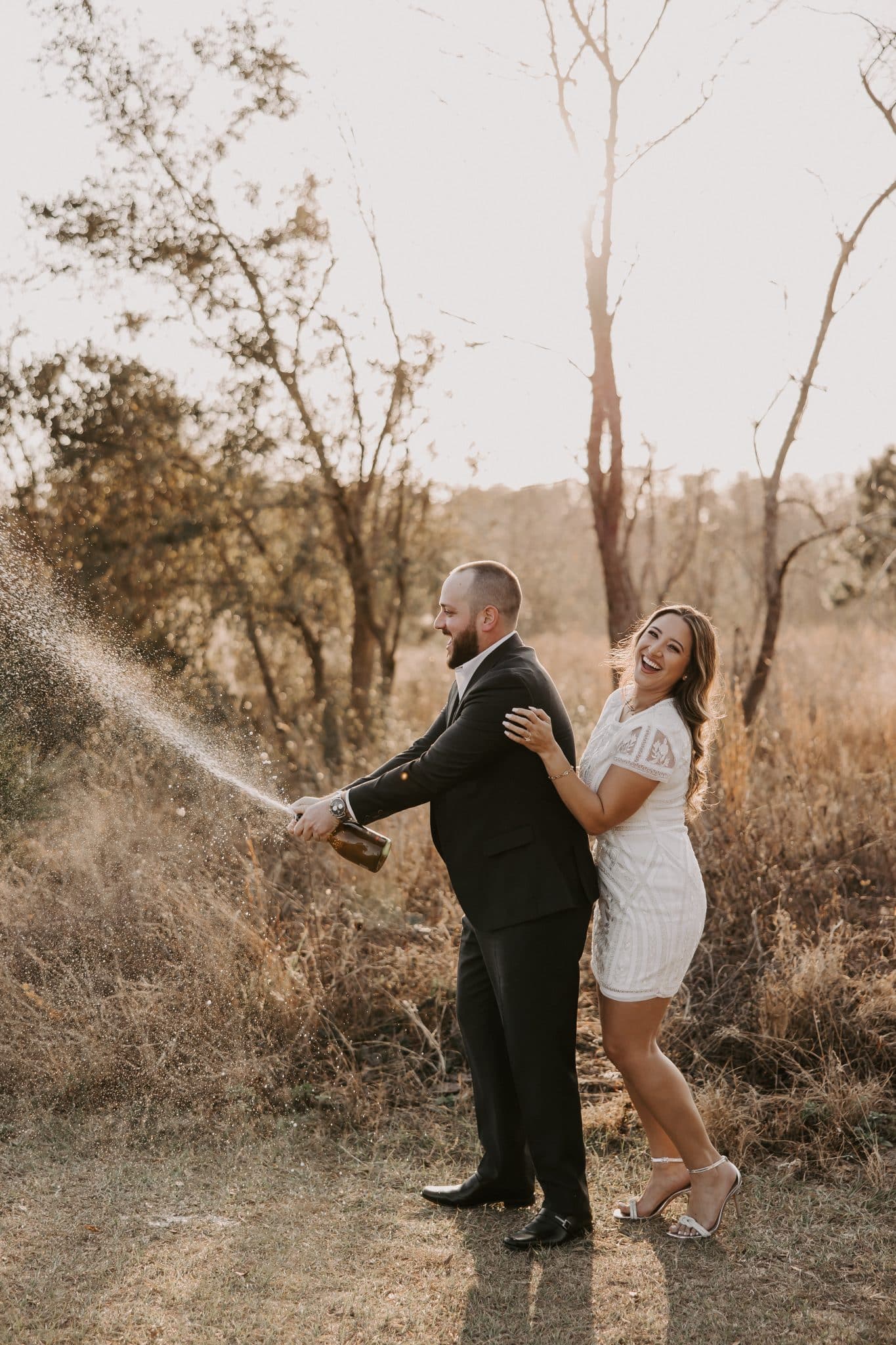 Newly engaged couple laughs together as Christian pops and sprays a bottle of champagne in celebration