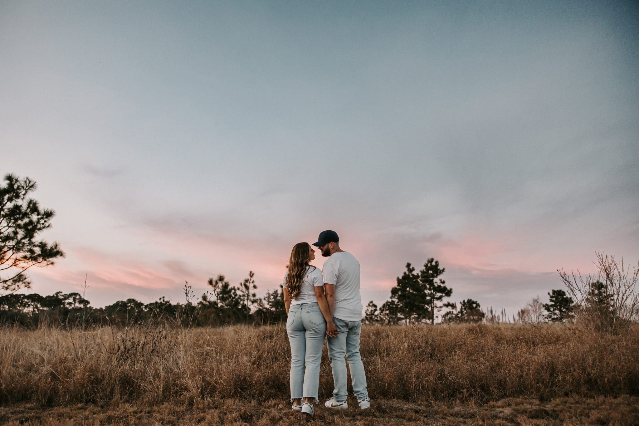 Couple stands in front of a pink and purple sunset in a field holding hands and gazing into each others eyes
