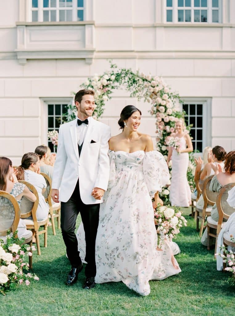bride wearing floral wedding gown and groom wearing white tuxedo dinner jacket from Carolyn Allen’s Bridals & Formals walking down the aisle