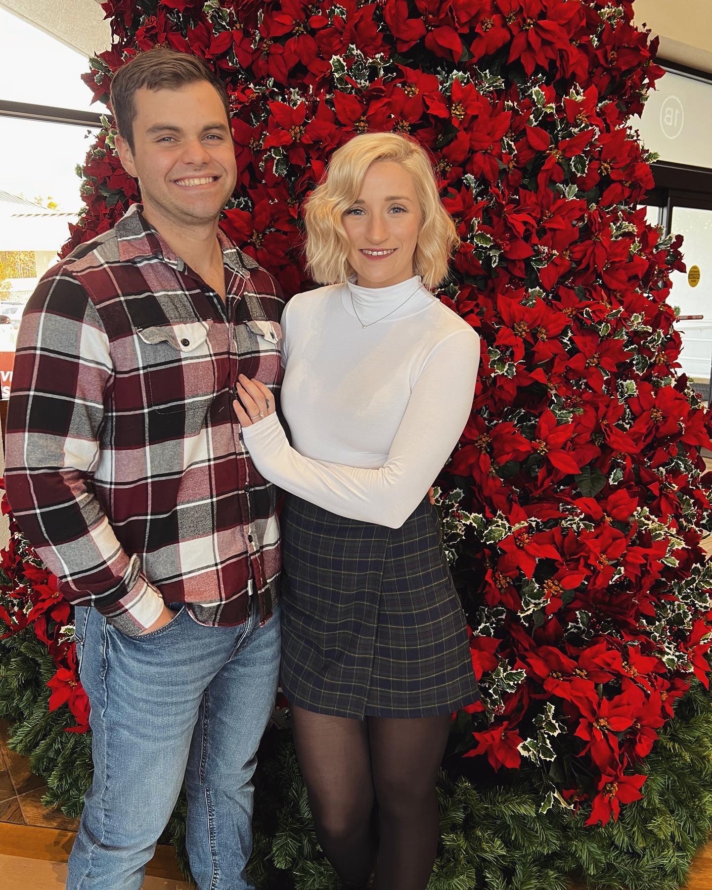 Couple poses for a photo together in front of a red Poinsette tree