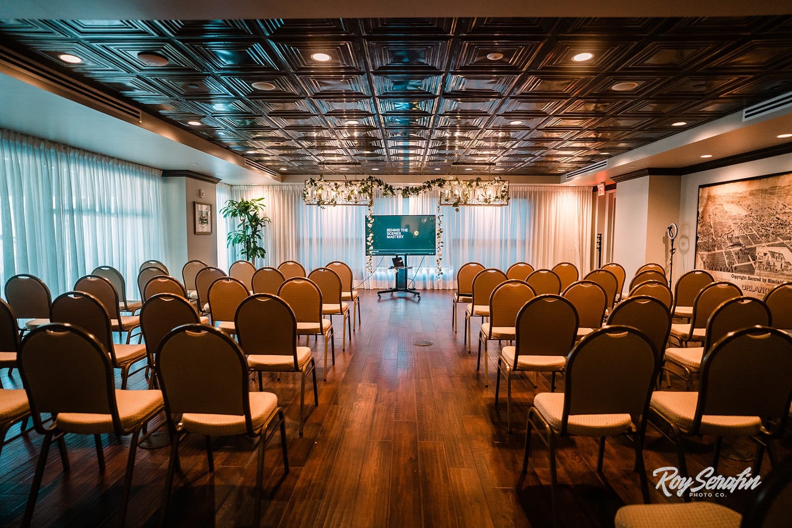 room setup with chairs facing the front of the room where a presentation tv is on a stand and floral is hanging of the lights above it for a speaker