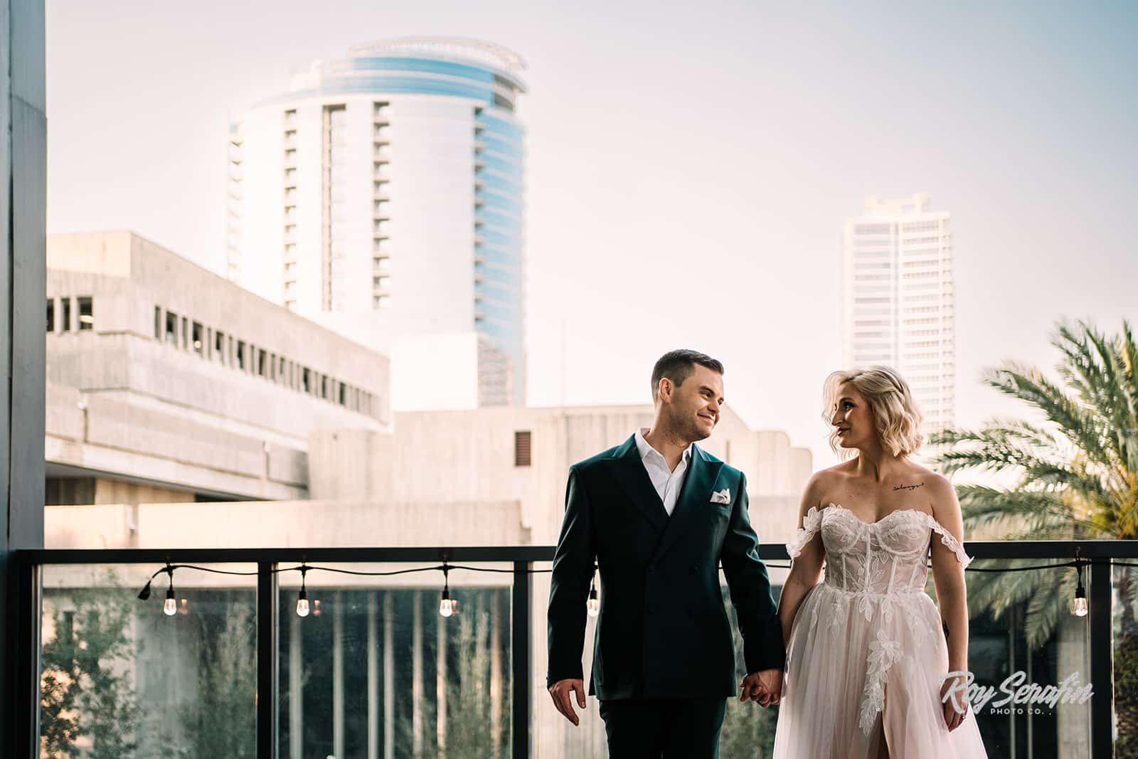 bride and groom hold hands and look at each other on wedding day outside in front of cityscape