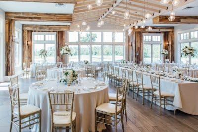 Elegant wedding reception hall at St. Johns Golf and Country Club in St. Augustine Florida featuring round tables with white linens, gold chiavari chairs, white floral centerpieces, rustic wood beam accents, and large windows overlooking the golf course greens