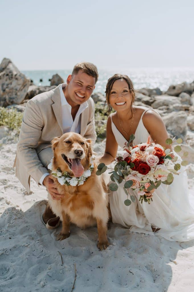 Bride and groom on the beach with their dog. The bride is holding a bouquet and their dog has a collar made of flowers.