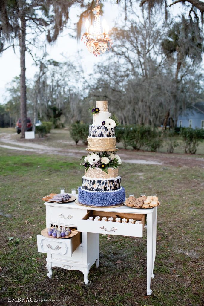 wedding cake in blue and gold set on desk in the outdoor reception area by Cake & Bake