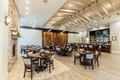 Elegant restaurant-style wedding reception dining room at St. Johns Golf and Country Club in St. Augustine Florida featuring wood beam ceiling accents, herringbone hardwood floors, stone fireplace, and full bar area with rustic wood wall backdrop