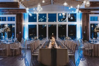 Elegant wedding reception hall at St. Johns Golf and Country Club in St. Augustine Florida featuring long head table and round guest tables with white linens candlelight centerpieces and warm pendant lighting against floor to ceiling windows with blue uplighting