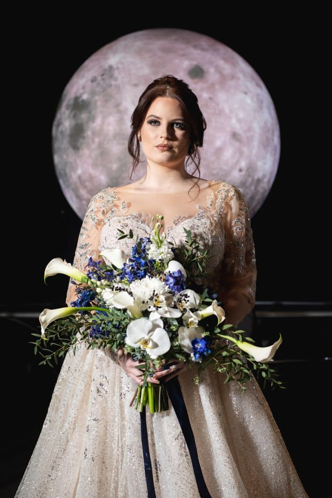 bride standing against a full moon backdrop with stunning bouquet of lilies and greenery from Dream Design Florist