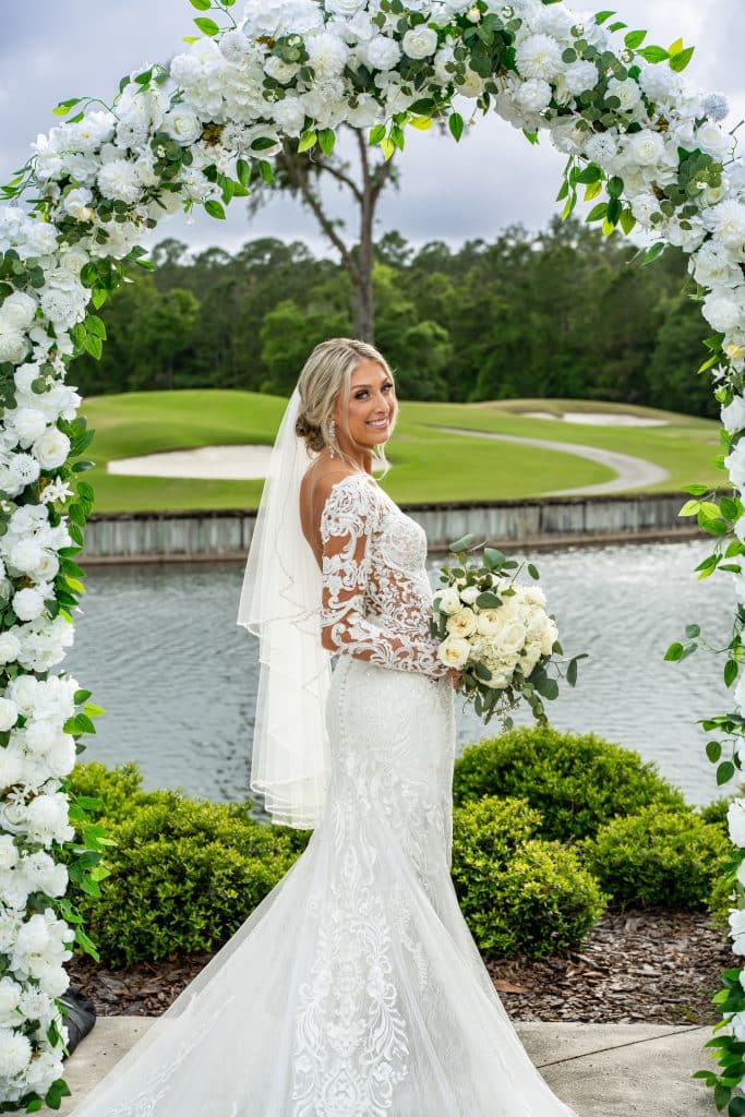 bride poses under arch with white flowers and golf course as the backdrop at St. Johns Golf & Country Club