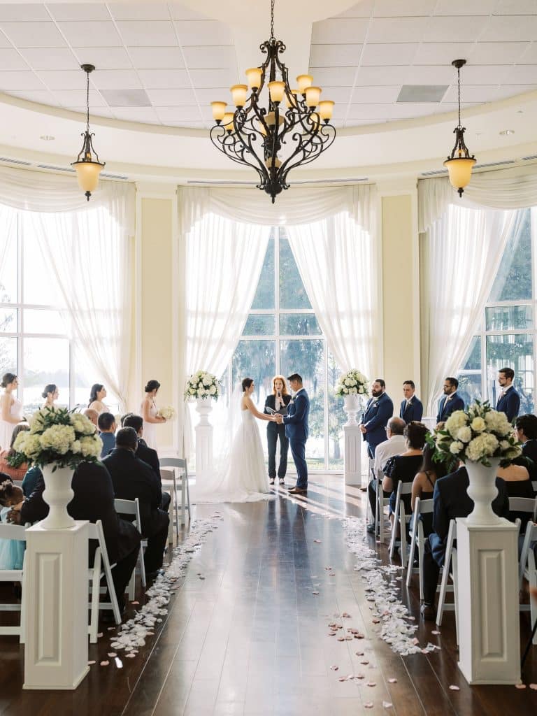bride and groom saying vows at the end of an aisle in a ballroom silhouetted against tall windows