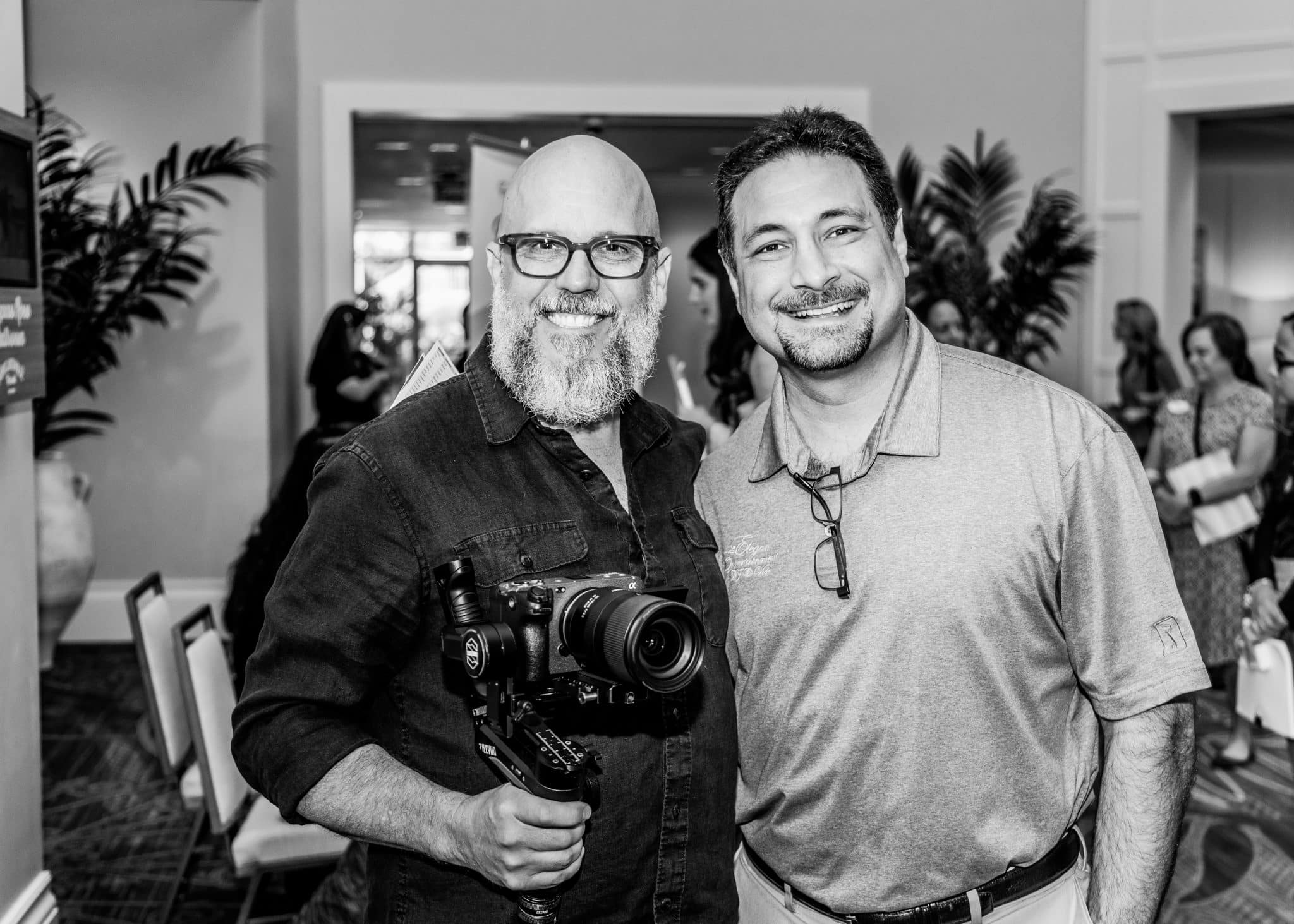 black and white image of two men standing together smiling for a picture while one holds a camera