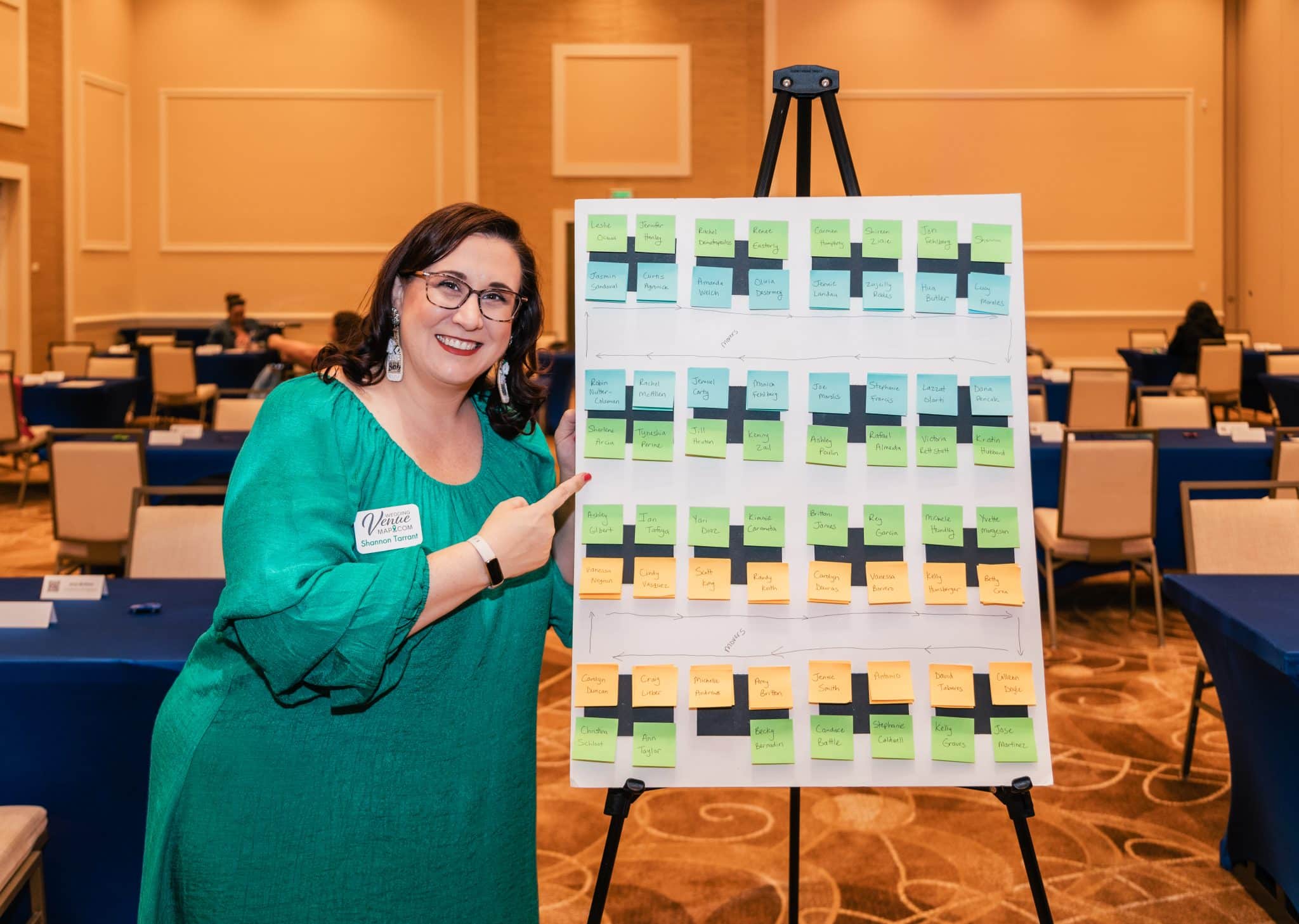 woman wearing green dress stands next to seating chart pointing to it and smiling