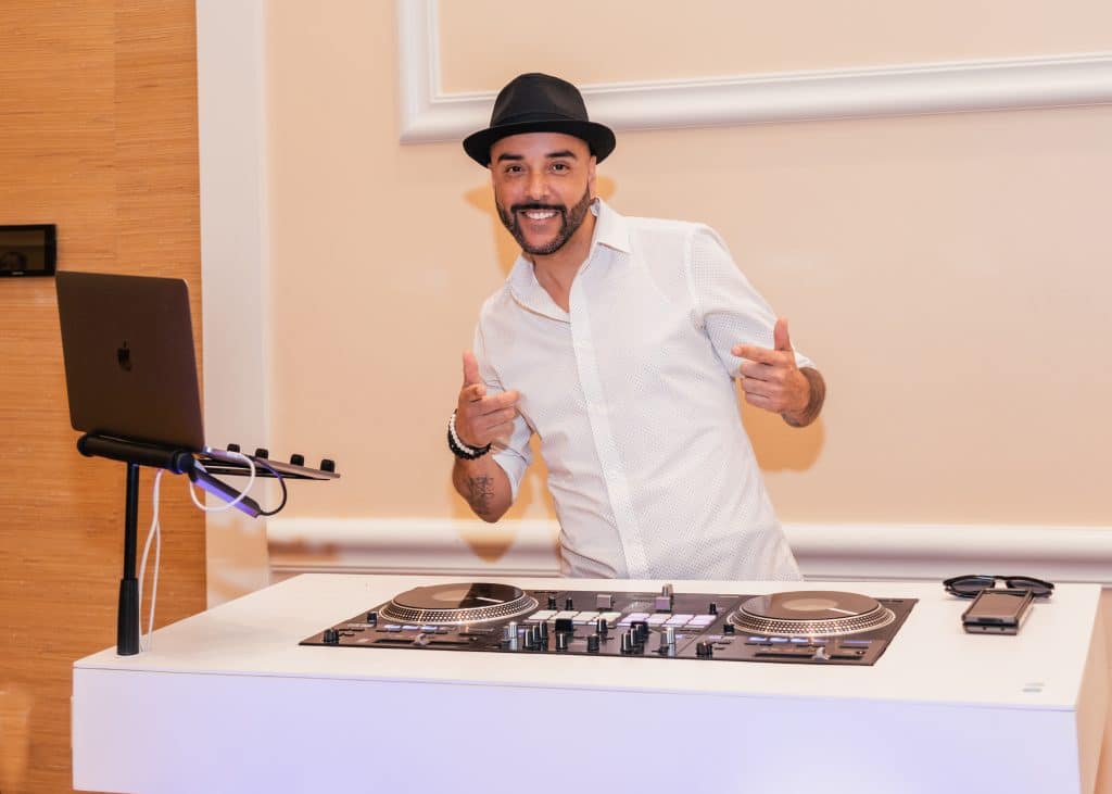 DJ stands behind his facade smiling with fedora hat on and white button up shirt