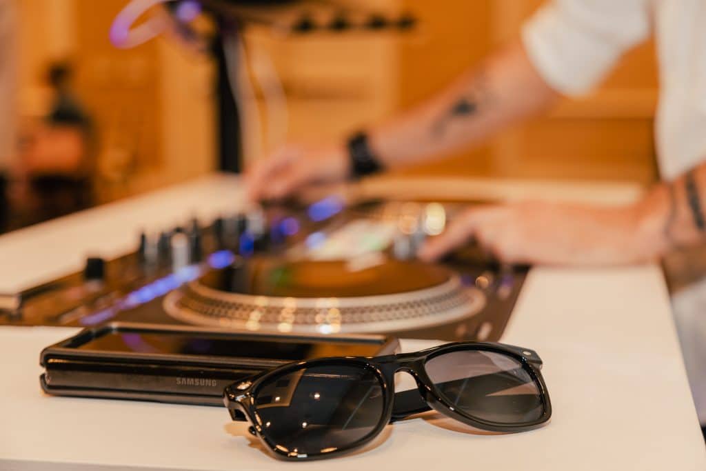 close up image of sunglasses sitting next to dj's mixing table while he uses it in the background