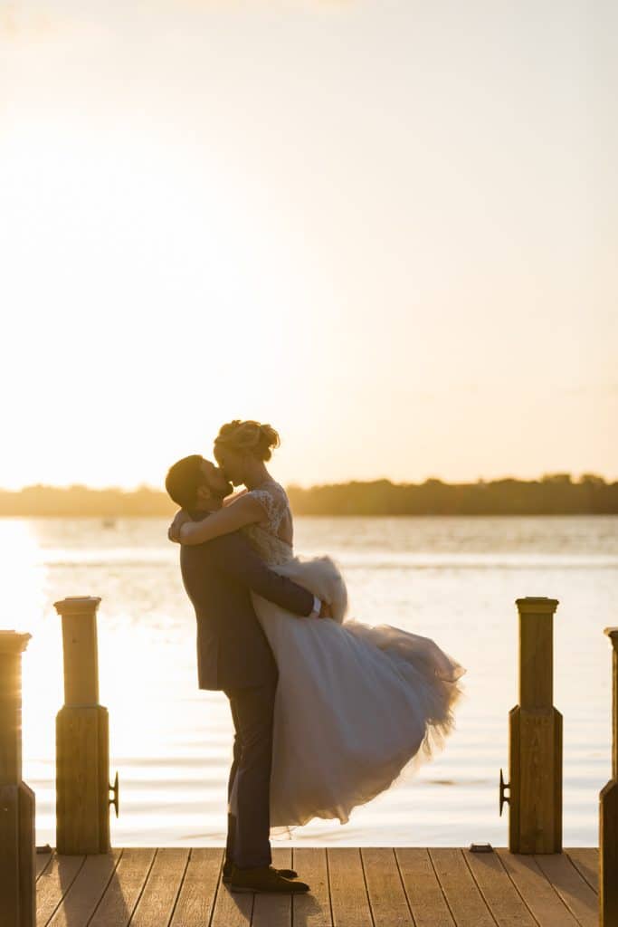 groom lifting bride for a kiss on a pier at sunset photographed by Ashley Jane Photography
