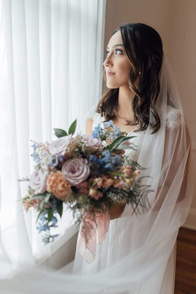 bride with mixed floral bouquet by Dream Designs Florist looking out window wrapped in her veil