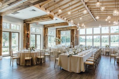 Elegant wedding reception hall at St. Johns Golf and Country Club in St. Augustine Florida featuring rustic wood beam ceilings with hanging globe pendant lights, long banquet tables and round tables draped in ivory linens with white floral centerpieces, gold chiavari chairs, and floor-to-ceiling windows overlooking lush green golf course grounds