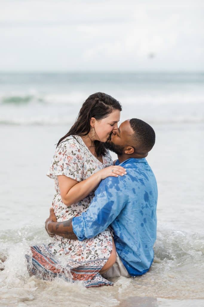 couple getting engaged and sharing a kiss sitting at the beach photographed by Ashley Jane Photography