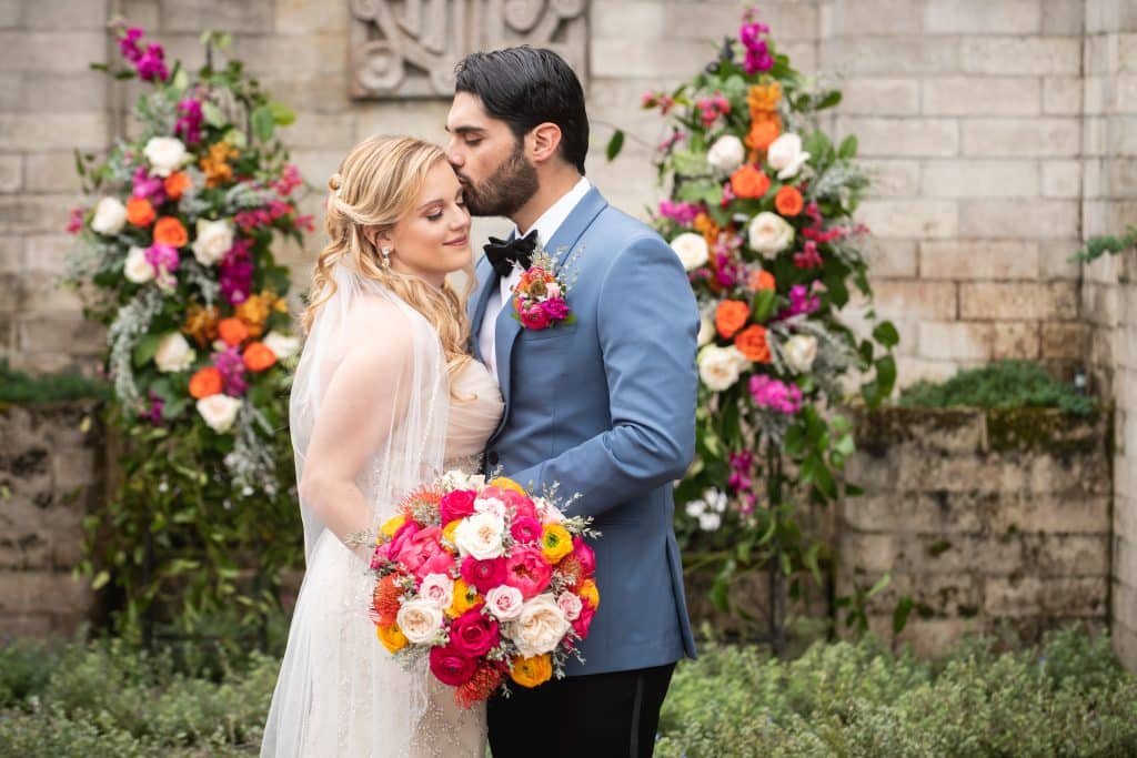 groom kissing bride as she holds her bouquet of colorful gerbera daisies and floral pillars by Dream Designs Florist behind them