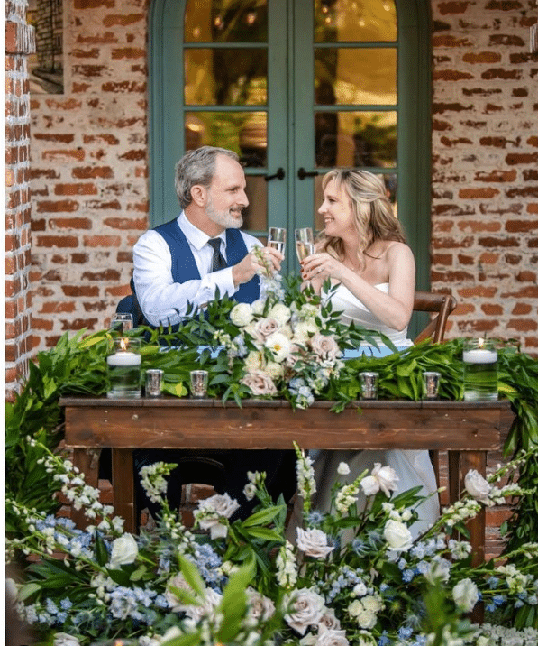 Couple toasting their marriage at the rustic wooden head table, outdoors, with a large floral bouquet on the table and in the foreground. The backdrop is exposed brick building with french doors, in Orlando, FL