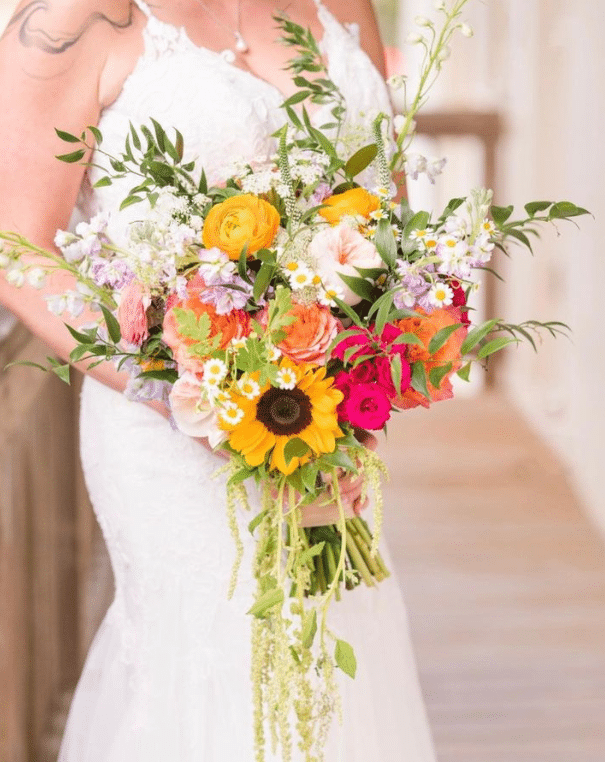Large floral bouquet with sunflowers, yellow, orange and pink flowers, held by a bride in a white gown, by Laynie Botanicals
