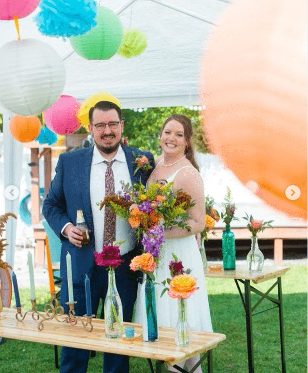 Happy couple standing together under a tent with paper lanterns hanging above. Floral arrangements on the table and the bride holding a vibrant bouquet by Laynie Botanicals