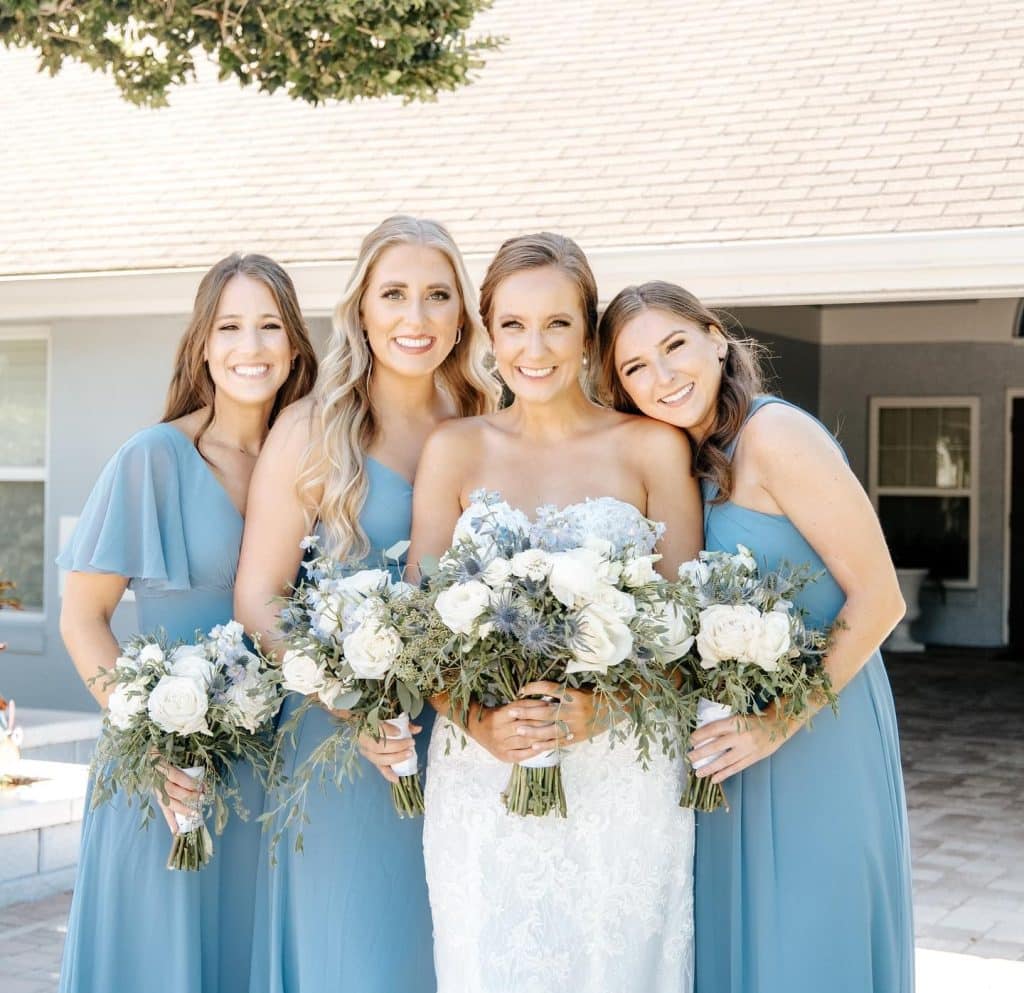 Wedding party posed shot wearing light blue dresses with the bride in a strapless white dress, all holding matching white floral bouquets in Orlando, FL, by Laynie Botanicals