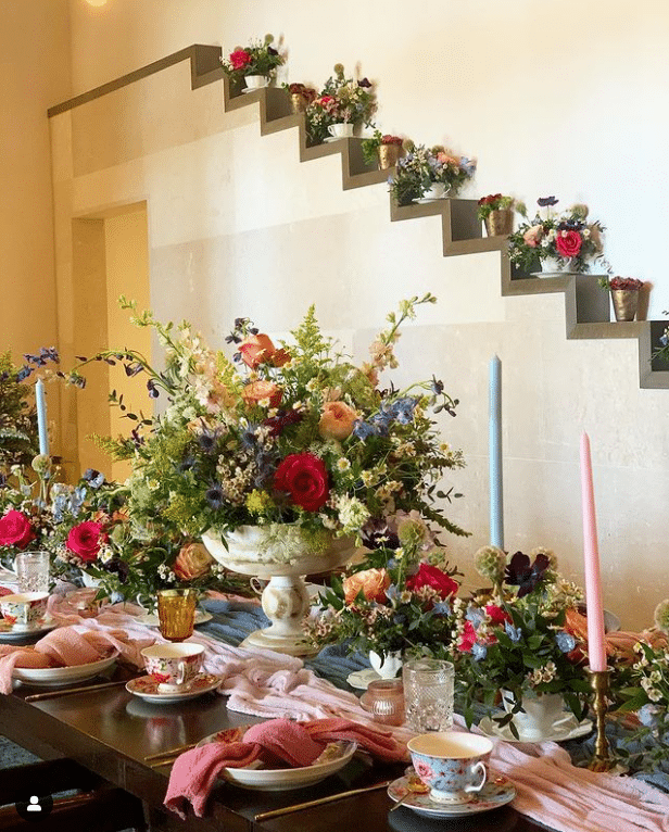 Large arrangement with bright vibrant colors in the center of a table, pink table runner, smaller table arrangements all along the table, background has an ascending wall design with a floral arrangement on each step